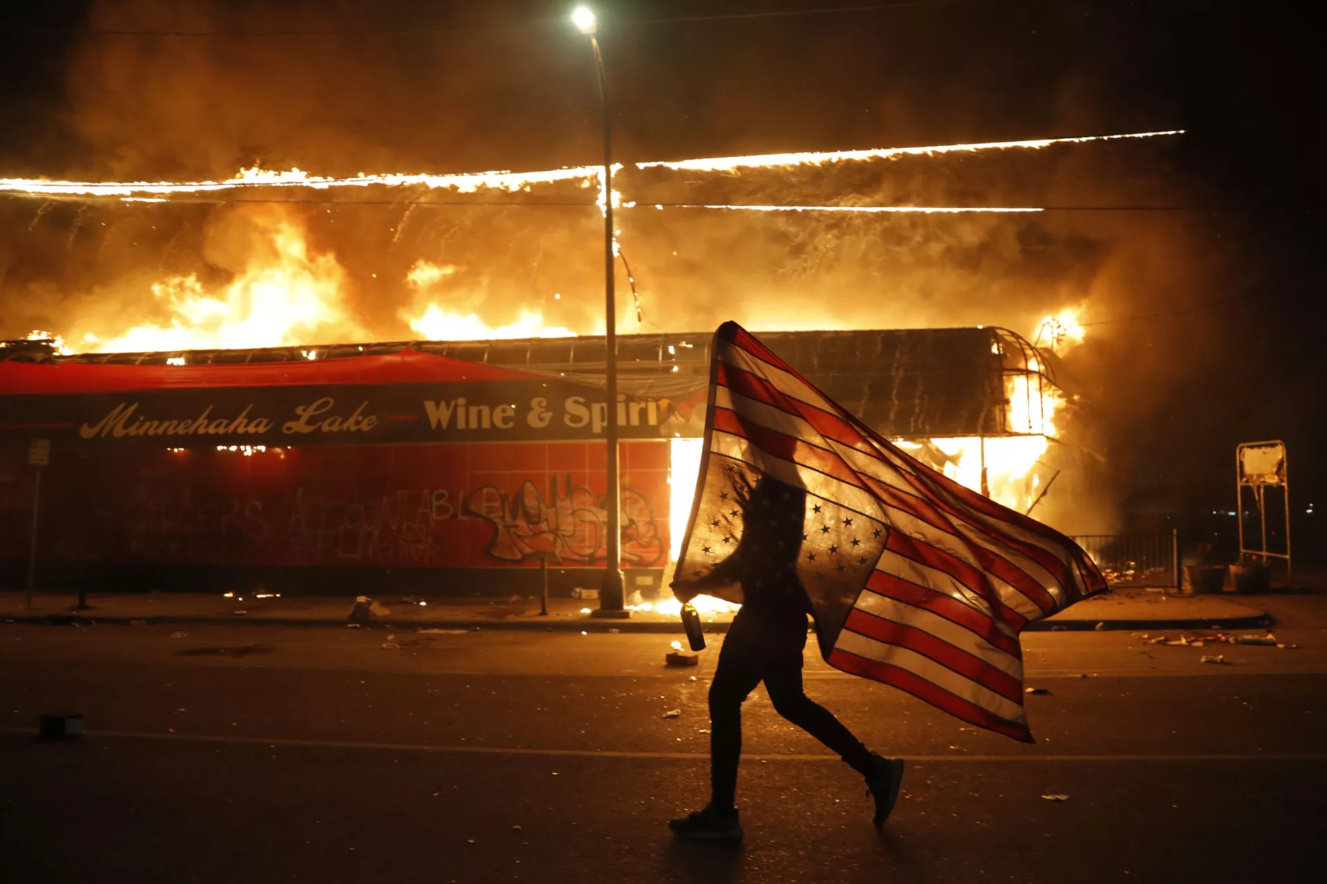 Un manifestante porta la bandera de EEUU boca abajo como símbolo de peligro extremo que vive el país frente un edificio en llamas en Minneapolis (estado de Minnesota), durante las protestas causadas por la muerte de George Floyd, el 28 de mayo del 2020 - Sputnik Mundo, 1920, 28.06.2021