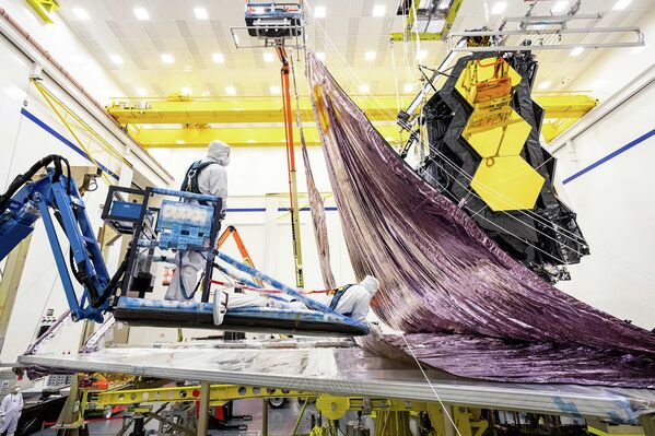 Tomada en febrero de 2021, esta imagen muestra el parasol del telescopio espacial James Webb siendo plegado y embalado por ingenieros y técnicos de Northrop Grumman. El parasol tiene una estructura de cinco capas en forma de diamante del tamaño de una pista de tenis, y ha sido especialmente diseñado para plegarse alrededor de los dos lados del telescopio, y caber dentro de los límites del cohete Ariane 5. Completamente desplegado, mide 21 metros por 14 metros, pero dentro del cohete (de 17 metros de largo y 5,4 metros de diámetro) ocupará un área muy reducida. - Sputnik Mundo