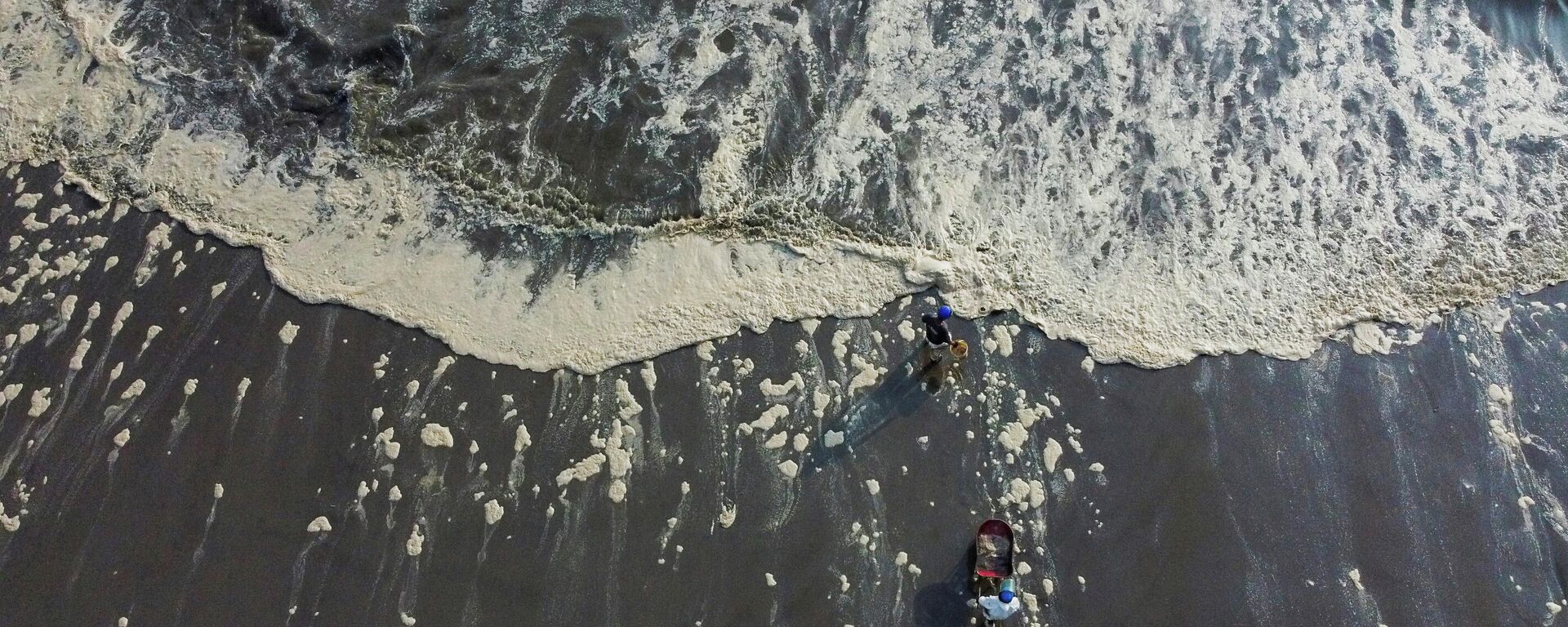 Unos trabajadores caminan entre la espuma en una orilla afectada por un vertido de petróleo causado por olas anómalas, provocadas por una enorme erupción volcánica submarina en Tonga, frente a la costa de Lima, en Ventanilla, Perú - Sputnik Mundo, 1920, 22.01.2022