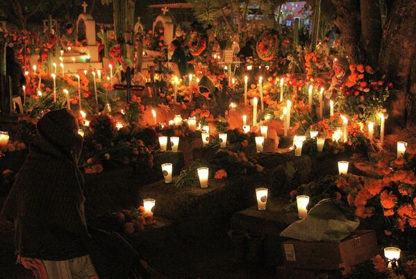 Una parte importante de la fiesta son los altares u ofrendas. En el Día de los Muertos, los familiares de los fallecidos preparan composiciones especiales que incluyen retratos de ellos, objetos importantes para ellos en vida y sus golosinas favoritas. En los cementerios, las familias hacen un pícnic en el que cenan junto a las tumbas de sus seres queridos.En la foto: velas del Día de los Muertos en el cementerio de Santa María Atzompa, México. - Sputnik Mundo