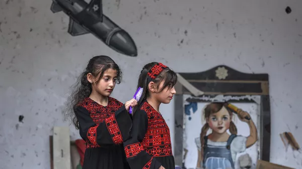 Una niña palestina cepilla el pelo de otra delante de un mural durante una exposición titulada 'La ocupación mata a la infancia' en Gaza, Palestina. - Sputnik Mundo