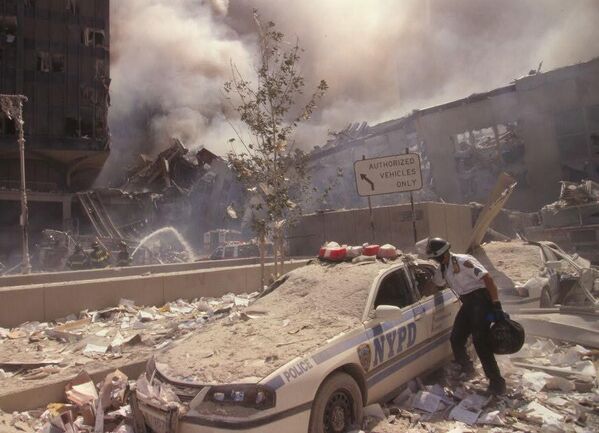 Un trabajador de rescate sube a un automóvil de la Policía de la ciudad de Nueva York cubierto de escombros mientras los bomberos rocían agua sobre las ruinas humeantes en el fondo. - Sputnik Mundo