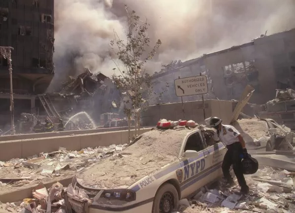 Un trabajador de rescate sube a un automóvil de la Policía de la ciudad de Nueva York cubierto de escombros mientras los bomberos rocían agua sobre las ruinas humeantes en el fondo. Un trabajador de rescate sube a un automóvil de la Policía de la ciudad de Nueva York cubierto de escombros mientras los bomberos rocían agua sobre las ruinas humeantes en el fondo. - Sputnik Mundo