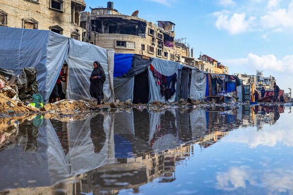 Palestinos desplazados caminando alrededor de un charco frente a edificios destruidos y tiendas en Jan Yunis, en el sur de la Franja de Gaza. - Sputnik Mundo