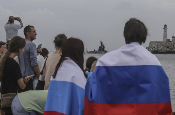 Los marinos rusos cumplirán un programa de actividades que incluye visitas de cortesía al jefe de la Marina de Guerra Revolucionaria y a la gobernadora de La Habana.En la foto: varias personas con banderas rusas observan la llegada del submarino nuclear ruso Kazan al puerto de La Habana. - Sputnik Mundo