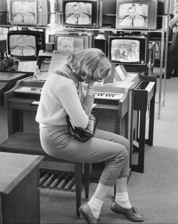 En el fondo de los televisores que retransmiten las noticias del asesinato del entonces presidente de EEUU, John F. Kennedy, una joven no identificada, con la mano cubriéndose la cara, está sentada en el supermercado Sears, Pensilvania, el 22 de noviembre de 1963. - Sputnik Mundo