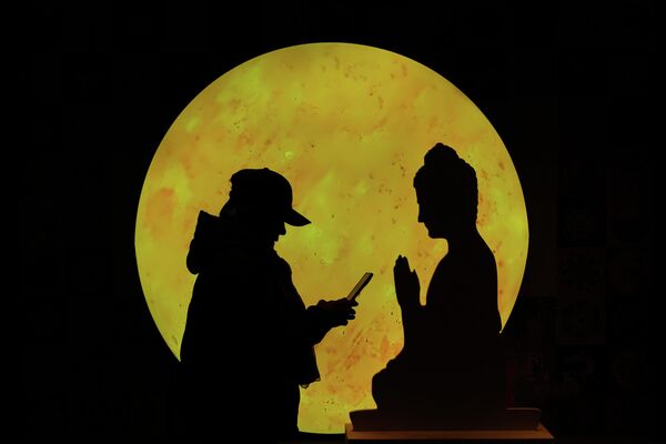 Una mujer consulta su celular frente a una figura de cartón de Buda con la luna de fondo, Pekín. - Sputnik Mundo