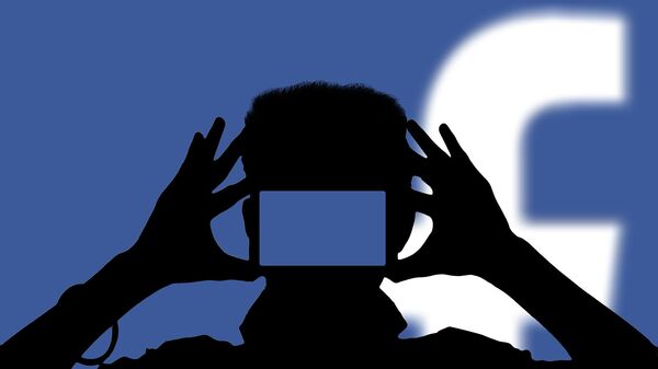 Una persona con un teléfono móvil con el logo de Facebook de fondo - Sputnik Mundo